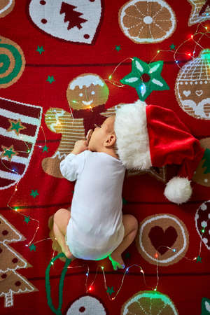 Little sleeping newborn baby, wearing Santa hat and white blank bodysuit, lies on red Christmas festive blanket with garland. Top above view, x-mas home indoorsの写真素材