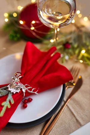 Traditional Christmas table place setting close up. Golden cutlery, linen napkin with deer ring, spruce branches and glass of wine. Holidays background. Selective focus, garland, bokeh.の写真素材