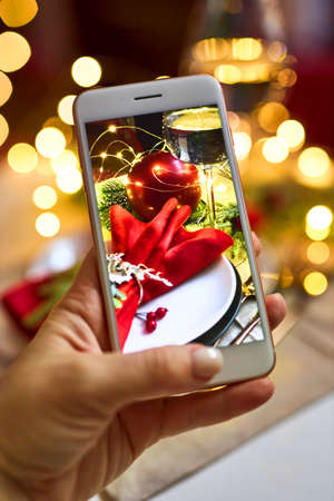Woman hand is taking pictures on phone the Christmas serving, festive table with lights, and using phone for blogging. Concept of making photos for social media posts.の写真素材