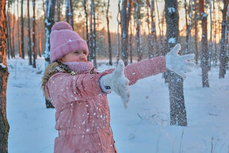 Child playing with snow in winter. Little girl in pink jacket and knitted hat catching snowflakes in winter park on Christmas. Kid play and jump in snowy forest. Snow ball fight for children.の写真素材