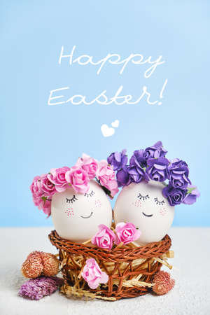 Two cuddling Easter eggs with sweet faces with floral spring wreaths lying in a cozy Easter basket. Creative postcard for Easter with copy spaceの写真素材