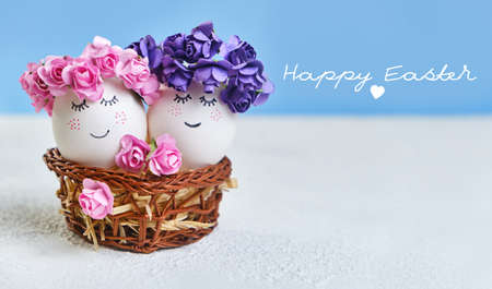 Two cuddling Easter eggs with sweet faces with floral spring wreaths lying in a cozy Easter basket. Creative postcard for Easter with copy spaceの写真素材