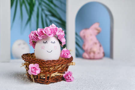 Natural white easter egg with drawn sweet face in floral wreaths in a wicker basket on the tropical beach with arches and a palm tree, in soft light. Happy Easter holiday card.の写真素材