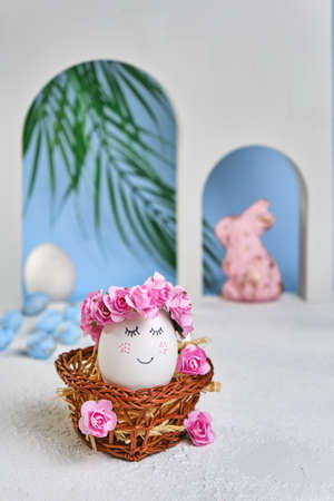 Natural white easter egg with drawn sweet face in floral wreaths in a wicker basket on the tropical beach with arches and a palm tree, in soft light. Happy Easter holiday card.の写真素材