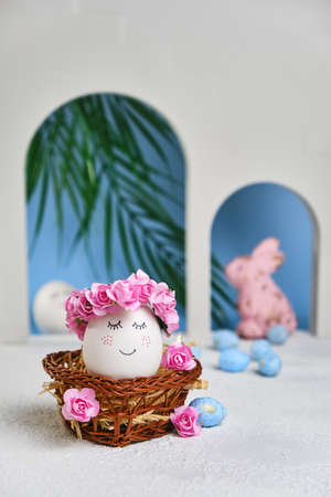 Natural white easter egg with drawn sweet face in floral wreaths in a wicker basket on the tropical beach with arches and a palm tree, in soft light. Happy Easter holiday card.の写真素材