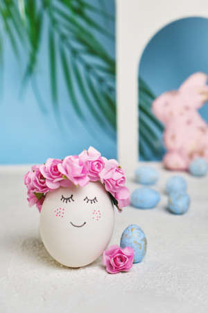 Natural white easter egg with drawn sweet face on the tropical beach with many little blue eggs, arches and a palm tree, in soft light. Happy Easter holiday card.の写真素材