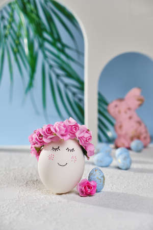 Natural white easter egg with drawn sweet face on the tropical beach with many little blue eggs, arches and a palm tree, in soft light. Happy Easter holiday card.の写真素材