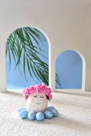 Natural white easter egg with drawn sweet face on the tropical beach with many little blue eggs, arches and a palm tree, in soft light. Happy Easter holiday card.の写真素材
