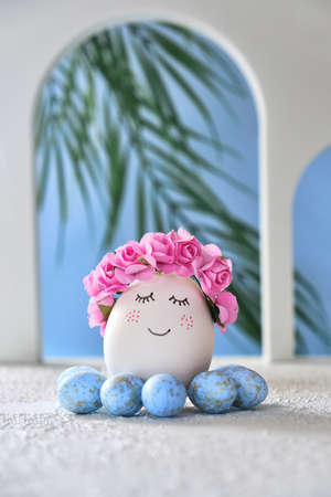 Natural white easter egg with drawn sweet face on the tropical beach with many little blue eggs, arches and a palm tree, in soft light. Happy Easter holiday card.の写真素材