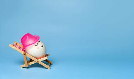 Creative funny composition with Easter egg wearing a hat sitting on deck chair on bright blue...