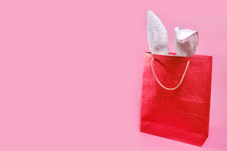 Easter bag with Easter decor. Bunny ears on a pink background with copy space. The concept of holiday shopping for Easter. Minimalistic wide banner with red gift bagの写真素材