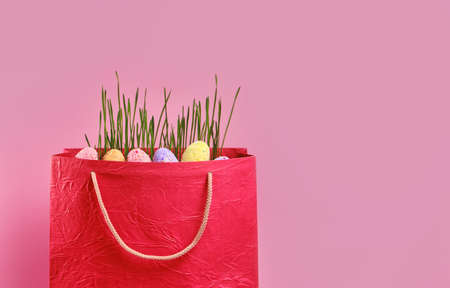 Green fresh juicy grass and multi-colored painted eggs in red shopping bag on pink background with copy spaceの写真素材
