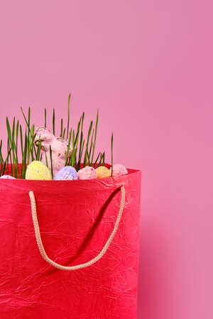 Green fresh juicy grass and multi-colored painted eggs and rabbit in red shopping bag on pink background with copy spaceの写真素材
