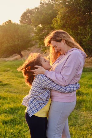 Side view on cheerful mother and daughter hugging each other, playing in the park on green grass in spring sunny day, happy family lifeの写真素材