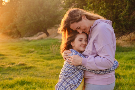 Side view on cheerful mother and daughter hugging each other, playing in the park on green grass in...