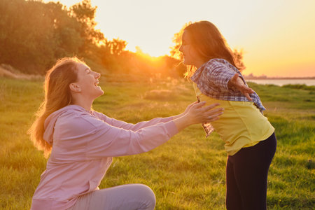 Side view on cheerful smiling mom and daughter looking to each other, playing in the park on green grass in spring sunny day, happy family lifeの写真素材