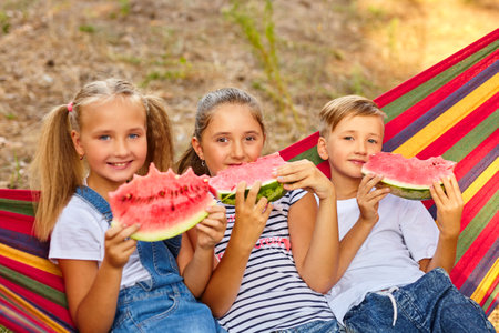three cheerful children eat watermelon and joke, outdoor, sitting on a colorful hammock. Summer fun and leisureの写真素材