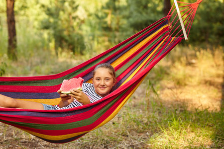 Cute little girl resting in a colored hammock in the forest and eating fresh watermelonの写真素材