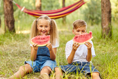 Happy Kids eating watermelon in the park. Kids eat fruits outdoors. Healthy snack for kids. Little girl and boy playing in the forest biting a slice of water melonの写真素材