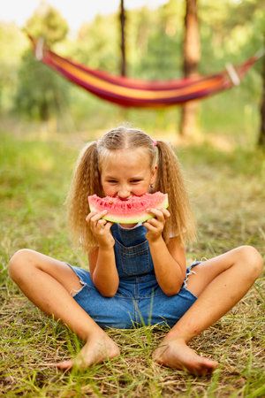Funny girl eating watermelon in the park. Kid eat fruits outdoors. Healthy snack for kids. Little girl playing in the forest biting a slice of water melonの写真素材