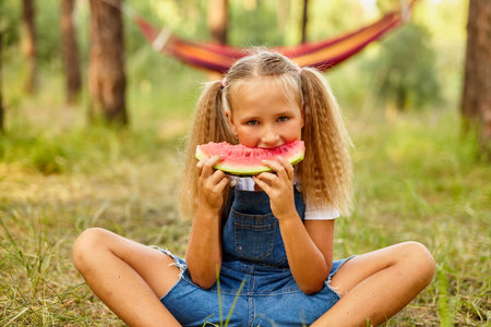 Funny girl eating watermelon in the park. Kid eat fruits outdoors. Healthy snack for kids. Little girl playing in the forest biting a slice of water melonの写真素材