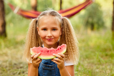 Funny girl eating watermelon in the park. Kid eat fruits outdoors. Healthy snack for kids. Little girl playing in the forest biting a slice of water melonの写真素材