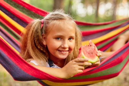 Cute little girl resting in a colored hammock in the forest and eating fresh watermelonの写真素材