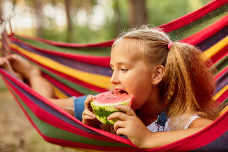 Cute little girl resting in a colored hammock in the forest and eating fresh watermelonの写真素材