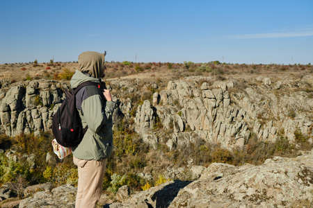 Hiker young man with backpack and trekking poles standing on edge of cliff and looking at the mountains in summer outdoorの写真素材