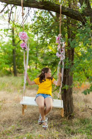Cute smiling girl in a yellow t-shirt having fun on a swing in tree forest. Sunny day. Summer outdoor activities for kidsの写真素材