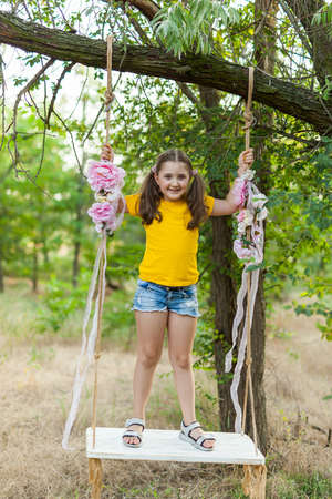 Cute smiling girl in a yellow t-shirt having fun on a swing in tree forest. Sunny day. Summer outdoor activities for kidsの写真素材