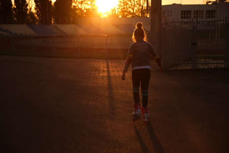 Little pretty happy funny girl on roller skates at stadium at sunset, learning to roller skate outdoors. outdoor activities for children. Active sport for preschool kid. sun glare, selective focusの写真素材