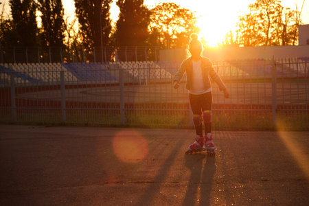 Little pretty happy funny girl on roller skates at stadium at sunset, learning to roller skate outdoors. outdoor activities for children. Active sport for preschool kid. sun glare, selective focusの写真素材