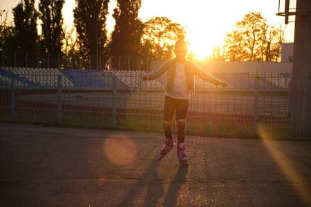 Little pretty happy funny girl on roller skates at stadium at sunset, learning to roller skate outdoors. outdoor activities for children. Active sport for preschool kid. sun glare, selective focusの写真素材