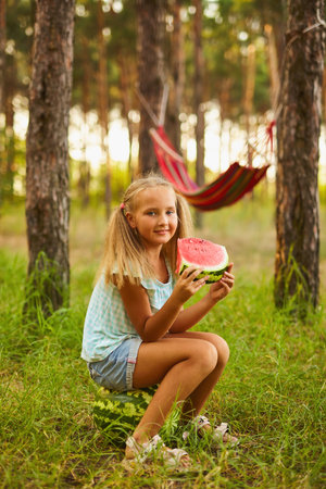 Summer season of watermelons. Picnic with a hammock in the forest. A cute little girl sits on a watermelon in the forest or garden and eating a sweet watermelonの写真素材