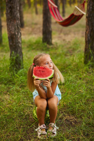 Summer season of watermelons. Picnic with a hammock in the forest. A cute little girl sits on a watermelon in the forest or garden and eating a sweet watermelonの写真素材