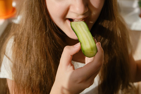 Kid bites cucumber, close-up, unrecognizable face. Girl eating cucumber at homeの写真素材