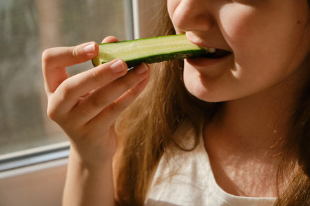 Kid bites cucumber, close-up, unrecognizable face. Girl eating cucumber at homeの写真素材