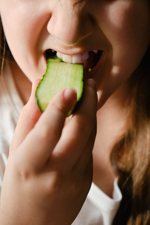 Kid bites cucumber, close-up, unrecognizable face. Girl eating cucumber at homeの写真素材
