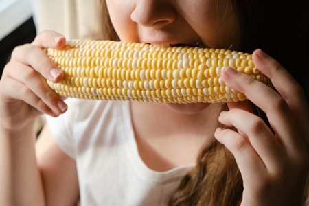 Close up child girl biting big vegetable corn at home, fresh vegetarian food, vitamins, healthy nutrition without GMOs. indoor. unrecognizable faceの写真素材