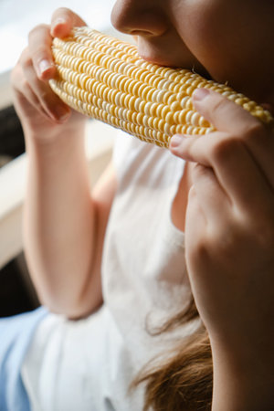 Close up child girl biting big vegetable corn at home, fresh vegetarian food, vitamins, healthy nutrition without GMOs. indoor. unrecognizable faceの写真素材