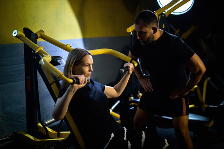Personal coach assisting sportswoman in using machine during gym workout in a dark gymの写真素材