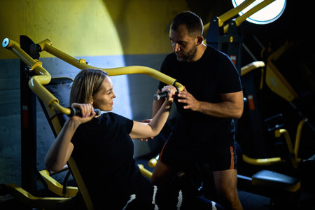 Personal coach assisting sportswoman in using machine during gym workout in a dark gymの写真素材