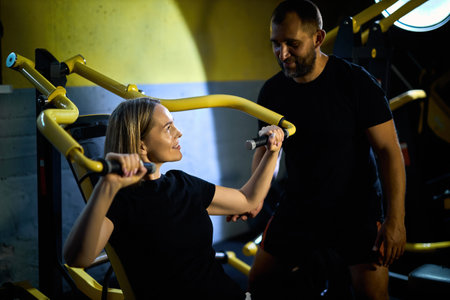 Personal coach assisting sportswoman in using machine during gym workout in a dark gymの写真素材