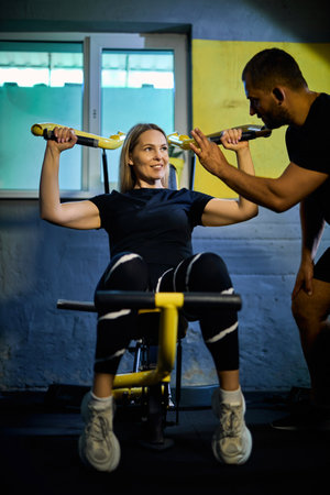 Personal coach assisting sportswoman in using machine during gym workout in a dark gymの写真素材