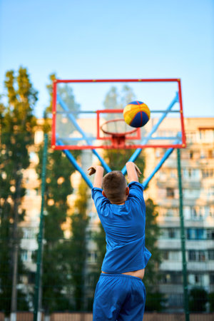 Joyful young sport boy kid playing basketball on the playground. Boy performs shot at red court ball game against the backdrop of urban buildings. Active lifestyle, training, professional sportsの写真素材