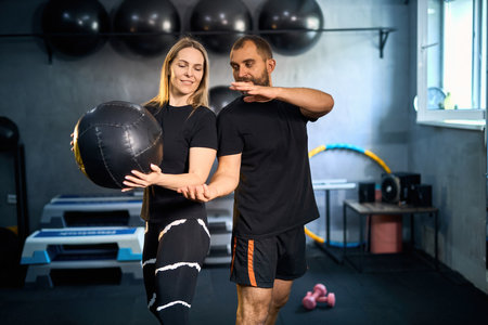Sporty couple exercising together in gym. Handsome man assisting beautiful girl while doing exercises with medicine ball in GYM.の写真素材