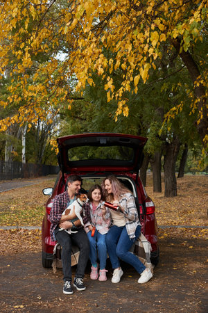 Happy parents with daughter and cute dog sitting in car trunk on autumn day. Long auto journey break.の写真素材