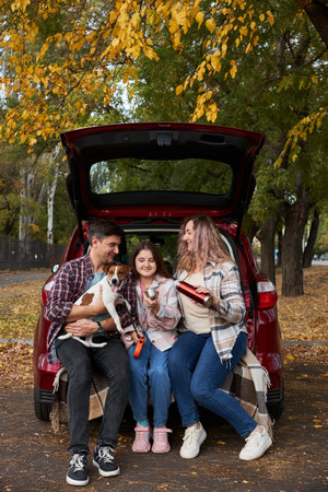 Happy parents with daughter and cute dog Jack Russel terrier sitting in car trunk on autumn day. Long auto journey break.の写真素材