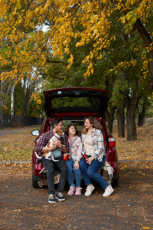 Happy parents with daughter and cute dog sitting in car trunk on autumn day. Long auto journey break.の写真素材